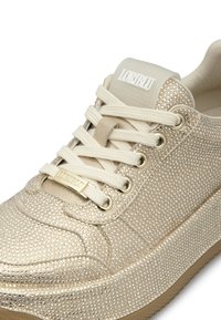Sneaker beige con tomaia testurizzata decorata con piccole borchie luccicanti, lacci crema e una piastra dorata. Logo visibile sulla linguetta.