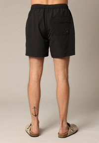 Shorts de bain noirs avec une taille élastique, poche arrière et ourlet plat. Le matériau semble léger et lisse. Sandales avec détail en liège.