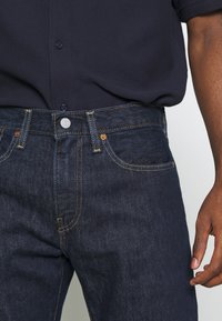Gros plan d'une personne portant un jean bleu foncé avec des coutures contrastées et une chemise bleu marine boutonnée et rentrée.