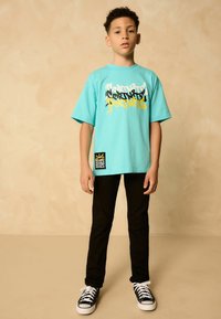 Tricou grafic turcoaz cu text multicolor, asortat cu pantaloni negri ajustați și pantofi sport negri cu glezna înaltă. Eticheta are un design cu coroană galbenă.