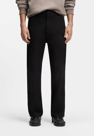 Hombre vistiendo pantalones negros rectos, zapatos tipo mocasín negros y una sudadera beige con las mangas remangadas.