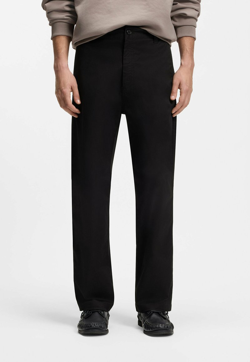 Homme portant un pantalon noir droit, des mocassins noirs et un sweat-shirt beige avec les manches retroussées.