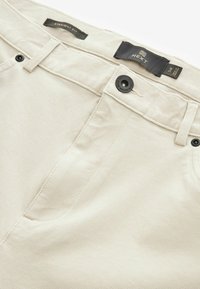 Pantalons beiges à coupe droite avec une texture lisse, des coutures visibles et un bouton noir. Présente une étiquette au niveau de la taille.