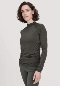 Langärmliges, dunkelgrünes Oberteil mit hohem Kragen, das an den Seiten gerafft ist und eine glatte Textur aufweist. Kombiniert mit schmal geschnittenen Hosen für einen eleganten Look.