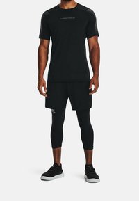 Hombre vistiendo camiseta de manga corta negra de Under Armour, pantalones cortos negros, mallas de compresión negras y zapatillas deportivas negras sobre fondo blanco.