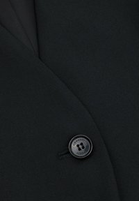 Czarna teksturowana tkanina marynarki z nachodzącymi się klapami, z czarnym guzikiem z wygrawerowanym napisem "ELEMENTY". Zbliżenie podkreśla detal i design.