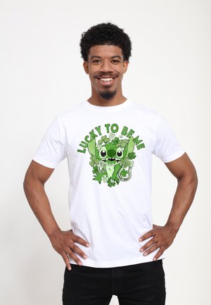 Lächelnder Mann mit lockigem Haar, der ein weißes T-Shirt trägt, auf dem eine grüne Cartoon-Figur von Kleeblättern und Hufeisen umgeben ist, mit dem Text "Glücklich, ich zu sein."