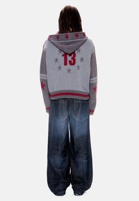 Άτομο με μακριά μαλλιά που φοράει γκρι φανέλα με κουκούλα, με κόκκινα αστέρια και τον αριθμό 13, συνδυασμένη με μπλε σκούρο τζιν με φαρδιά πόδια, με την πλάτη προς τα εμπρός.