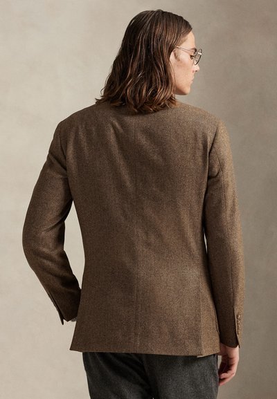 Άνδρας με μαλλιά μέχρι τους ώμους, φοράει γυαλιά και καφέ καρώ σακάκι, στέκεται με την πλάτη προς ένα απλό μπεζ φόντο.