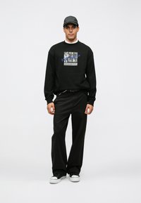 Felpa nera con stampa grafica, abbinata a pantaloni neri e sneakers nere con suola bianca. Il modello indossa un cappellino abbinato.