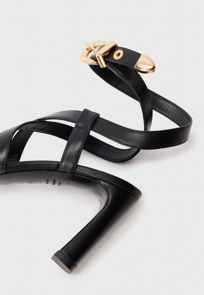 Zwarte leren hoge hak sandaal met gekruiste banden, een glanzende gouden gesp en een elegante stiletto hak, met een afgeronde neus en een gladde textuur.