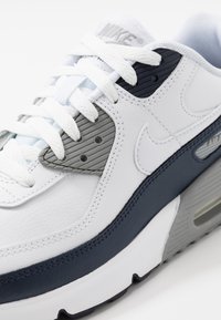 Zapatillas Nike Air Max con cuero blanco, acentos en azul marino y gris, cordones prominentes y marca en la lengüeta y el costado.