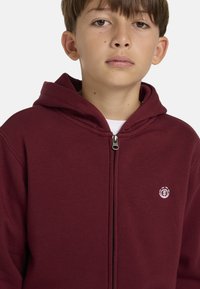 Sudadera con cremallera en color burdeos, con capucha y fabricada en un tejido suave. Presenta un pequeño logo en el lado izquierdo del pecho y una cremallera de metal.