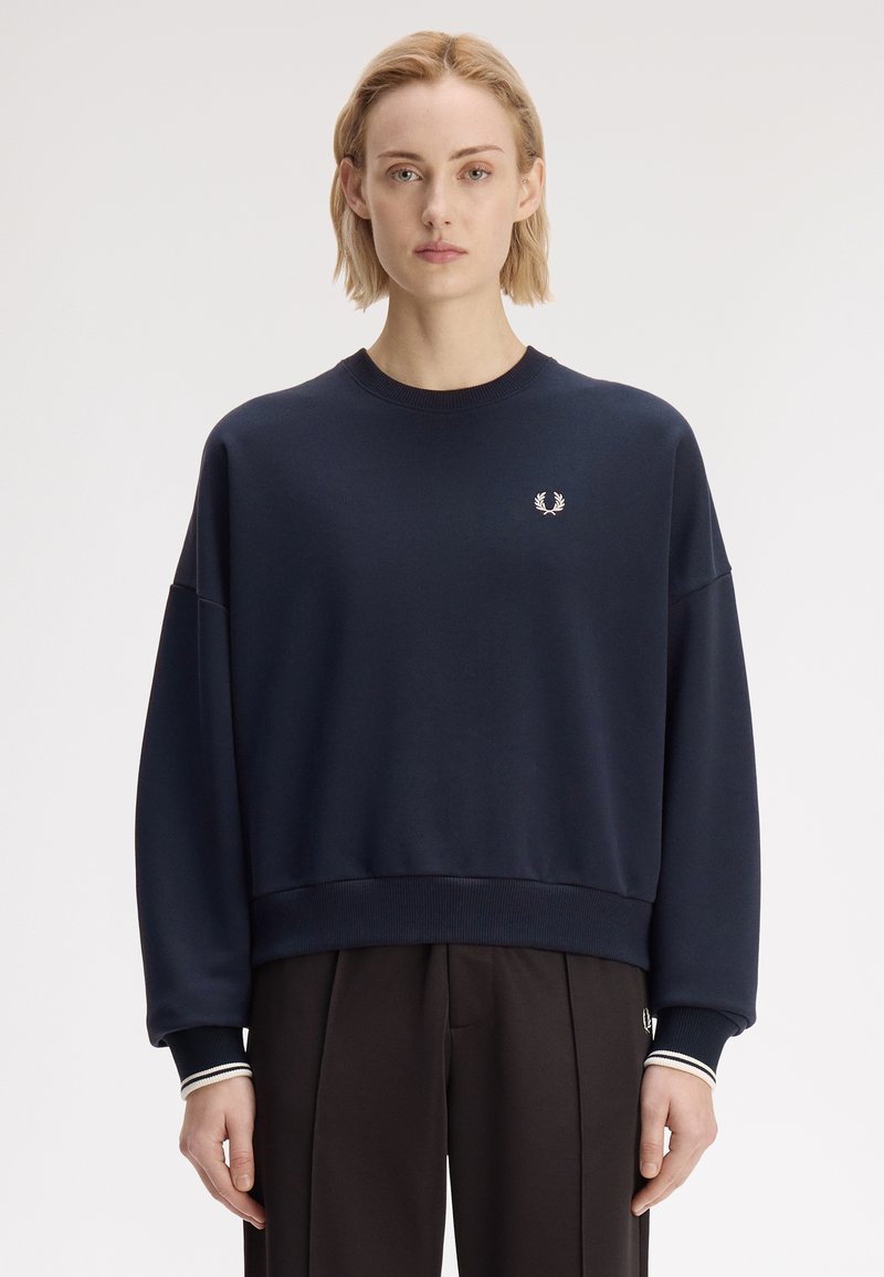Personne aux cheveux blonds courts portant un sweat-shirt bleu marine avec un petit logo de laurier blanc sur la poitrine et un pantalon noir.