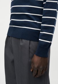 Pull rayé bleu marine avec des rayures blanches, poignets et ourlet côtelés, associé à un pantalon gris. Gros plan sur le tissu texturé du vêtement.