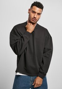 Hombre con cabello corto y rizado, vistiendo sudadera negra y jeans azules, posando con una mano cerca de su cuello sobre un fondo gris.