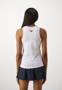 Top sportivo bianco con un design aderente, caratterizzato da un piccolo logo nero sulla parte superiore della schiena e una texture liscia. Indossato con una gonna svasata blu navy.