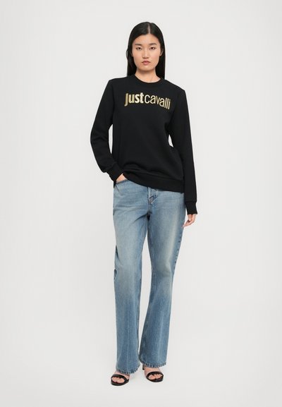 Femme debout dans un sweat-shirt noir avec le texte doré "Just Cavalli", un jean bleu clair et des talons ouverts noirs, sur fond blanc.