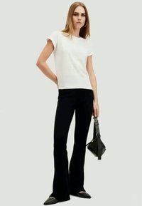 T-shirt branca de manga curta combinado com calças pretas de boca de sino. A modelo segura uma pequena mala preta e usa sapatos pretos planos. Design simples e ajustado.