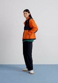 Giacca in fleece nera e arancione indossata sopra una maglietta verde, abbinata a pantaloni neri e sneakers bianche, su uno sfondo grigio chiaro.