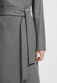 Cappotto di lana grigio con design avvolgente, dotato di una cintura coordinata e dettagli metallici alle estremità. Il tessuto presenta un aspetto morbido e strutturato.