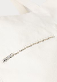 White canvas bag featuring a horizontal zippered pocket with a silver zipper, set against a textured fabric backdrop.