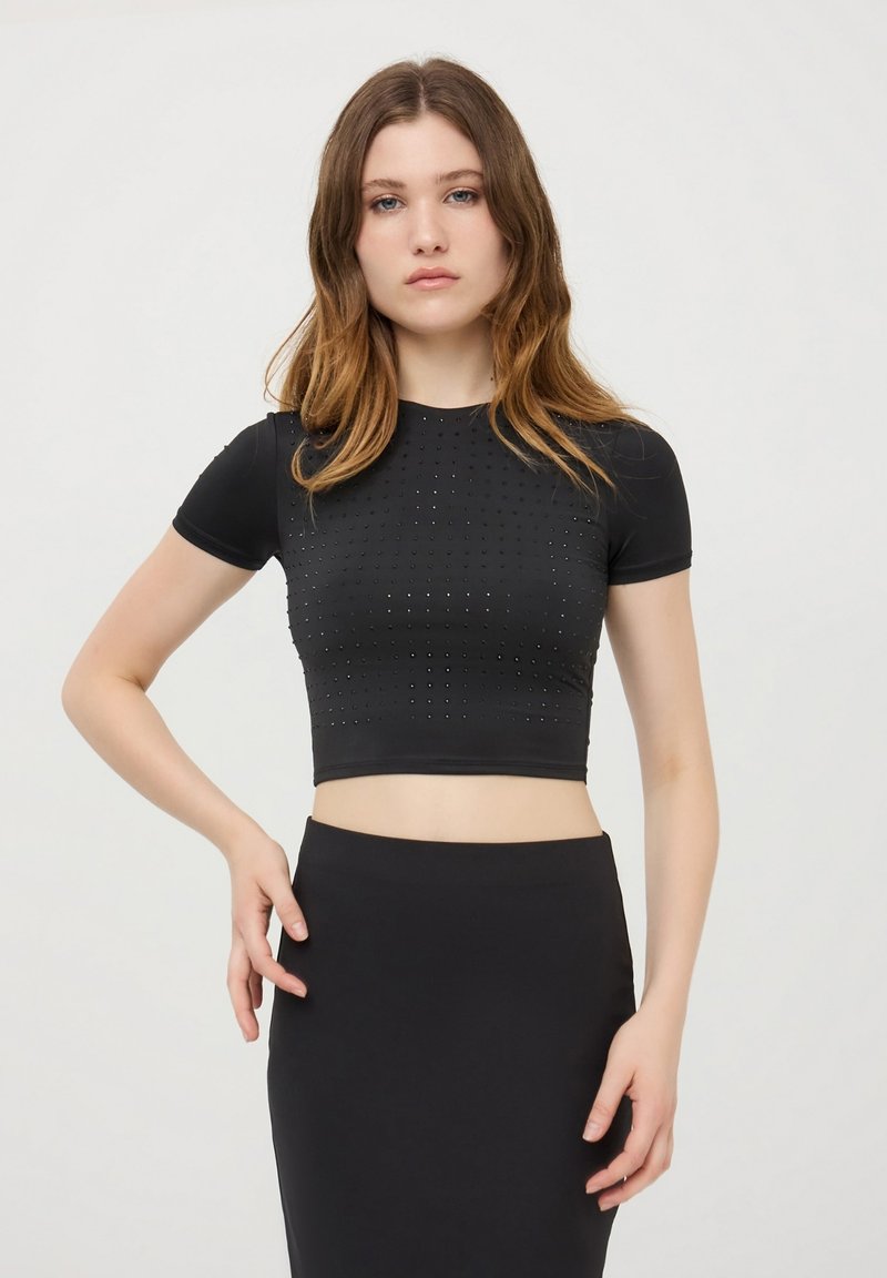 Cropped top nero realizzato in tessuto elasticizzato, con maniche corte e dettagli borchiati sul davanti. Il design è aderente e slanciato.