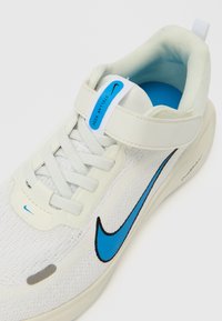 Fehér tornacipő kék Nike logóval, texturált felsőrésszel, fűzős záródással, tépőzáras pánttal és párnázott szegéllyel a kényelemért.