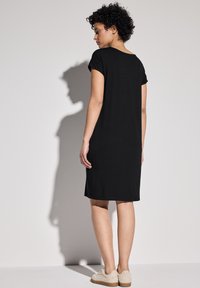 Svart T-shirt klänning med korta ärmar, rak passform, mjukt tyg, som når midjan på vaden, kombinerad med ljusa platta skor.