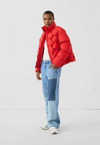 Rote Wattierte Jacke mit hohem Kragen und gesteppter Musterung, kombiniert mit hellblauen Patchwork-Denim-Jeans und weißen Sneakern.