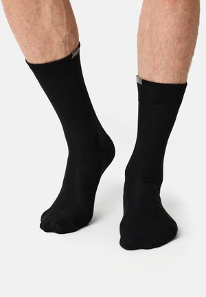 Uomo con calzini neri alla caviglia in piedi su una superficie bianca, mostrando entrambi i piedi con gambe inferiori e caviglie visibili.