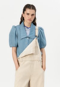 Camicia di denim con maniche a sbuffo, colletto grande; abbinata a salopette beige a righe con bretelle regolabili e tasche laterali.