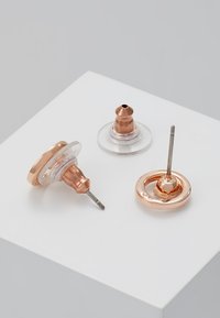 Brincos de studs em ouro rosa com suporte de plástico transparente, apresentando um design circular e um pino afunilado para um uso seguro.