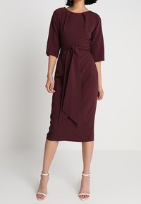 Robe portefeuille bordeaux avec une taille cintrée, des manches trois-quarts et un ourlet à hauteur des genoux. Texture lisse avec un détail de ceinture nouée à la taille.