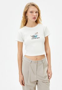 Haut court à côtes blanches avec une illustration imprimée de deux chats et le texte "LES CHATS SONT LIQUIDES." Associé à un pantalon beige clair.