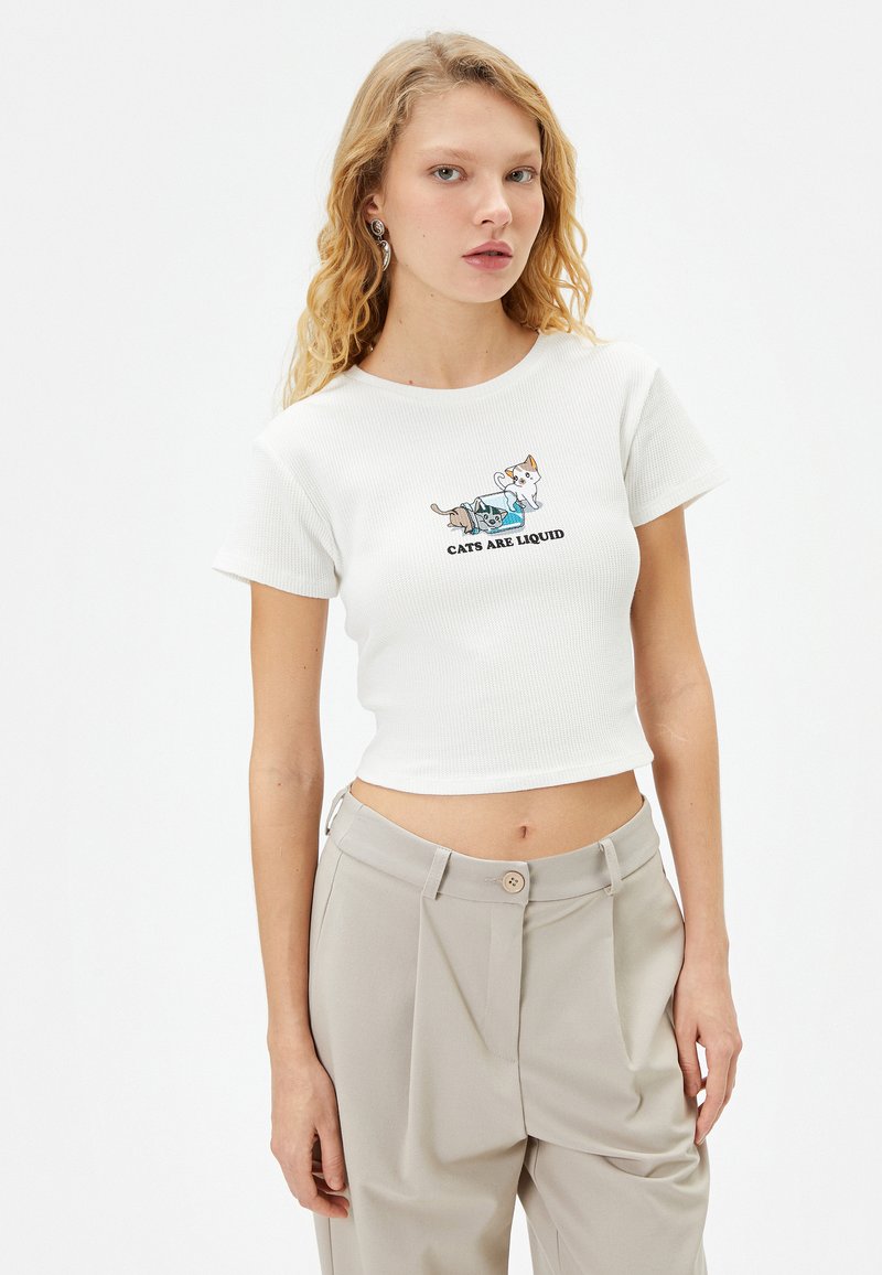 Haut court à côtes blanches avec une illustration imprimée de deux chats et le texte "LES CHATS SONT LIQUIDES." Associé à un pantalon beige clair.
