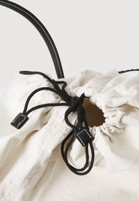 Canvas drawstring bag with black cord and accents, cream-colored fabric, gathered top, and decorative stitching on the drawstring ends.
