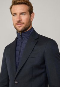 Blazer navy con una leggera texture sopra un giubbotto blu imbottito con colletto alto e chiusura con zip; tasca laterale sul blazer.