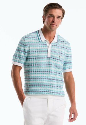 Homme portant un polo en maille à manches courtes bleu et vert à motifs, avec col et poignets blancs, associé à un pantalon blanc.