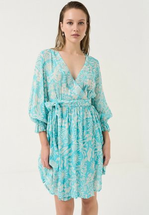 Kvinde iført en lyseblå blomsterprintet slå-om kjole med trekvartlange puffærmer og et bindebælte i taljen, stående mod en ensfarvet baggrund.