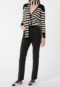 Cardigan noir et crème rayé avec un col en V, associé à un pantalon ajusté noir. La tenue comprend une écharpe rayée assortie et un accent en chaîne.