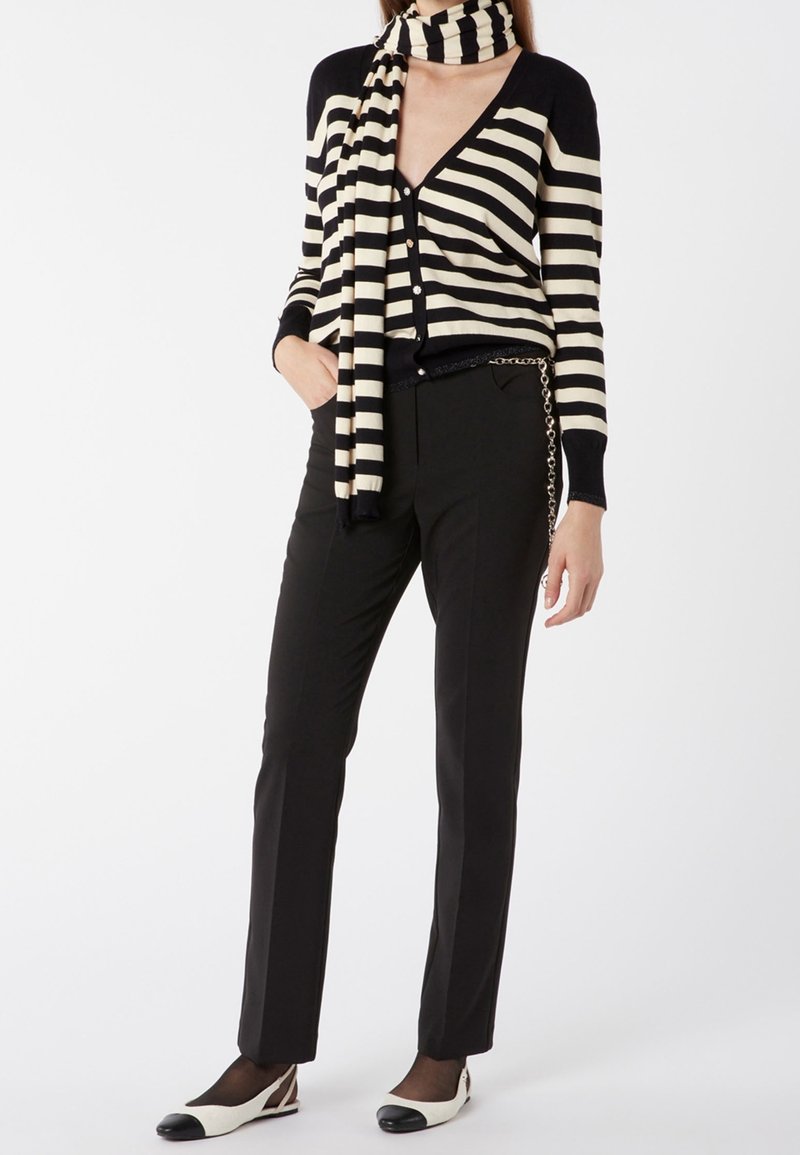 Cardigan noir et crème rayé avec un col en V, associé à un pantalon ajusté noir. La tenue comprend une écharpe rayée assortie et un accent en chaîne.