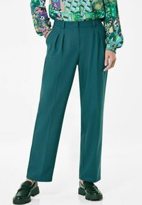 Teal wijde broek met plooien, gemaakt van een zachte stof. Gecombineerd met donkergroene platform instappers en een blouse met een kleurrijk patroon.