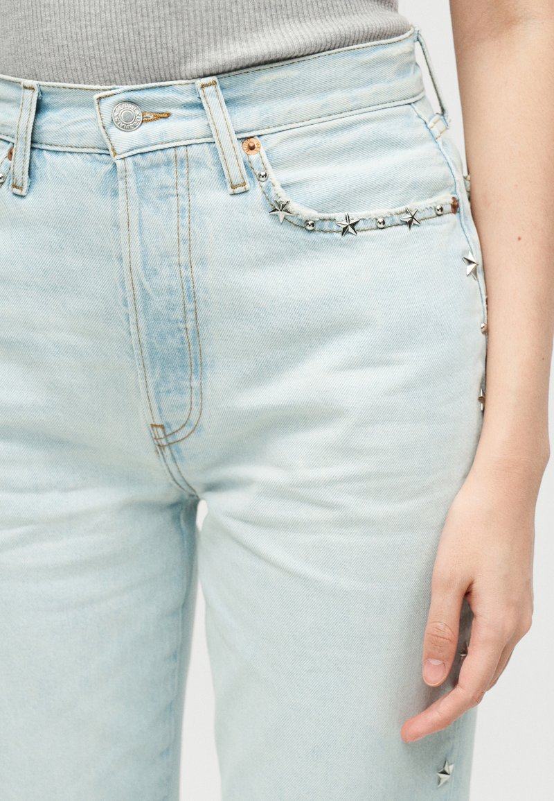 Jean en denim bleu clair avec des clous argentés en forme d’étoile sur les poches et l’extérieur de la cuisse, porté avec un haut beige à côtes.