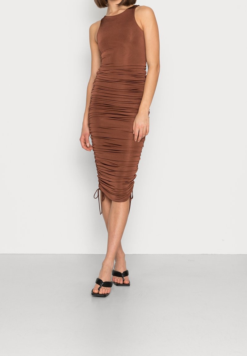 Žena v hnědé midi šatech bez rukávů se stažením po stranách, které jsou nastavitelné, v kombinaci s černými sandály na podpatku s otevřenou špičkou, stojící na šedé podlaze.