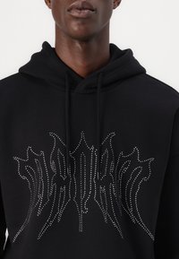 Black hoodie made of smooth fabric with a front design featuring silver studs in a flame-like pattern. Includes a drawstring hood.