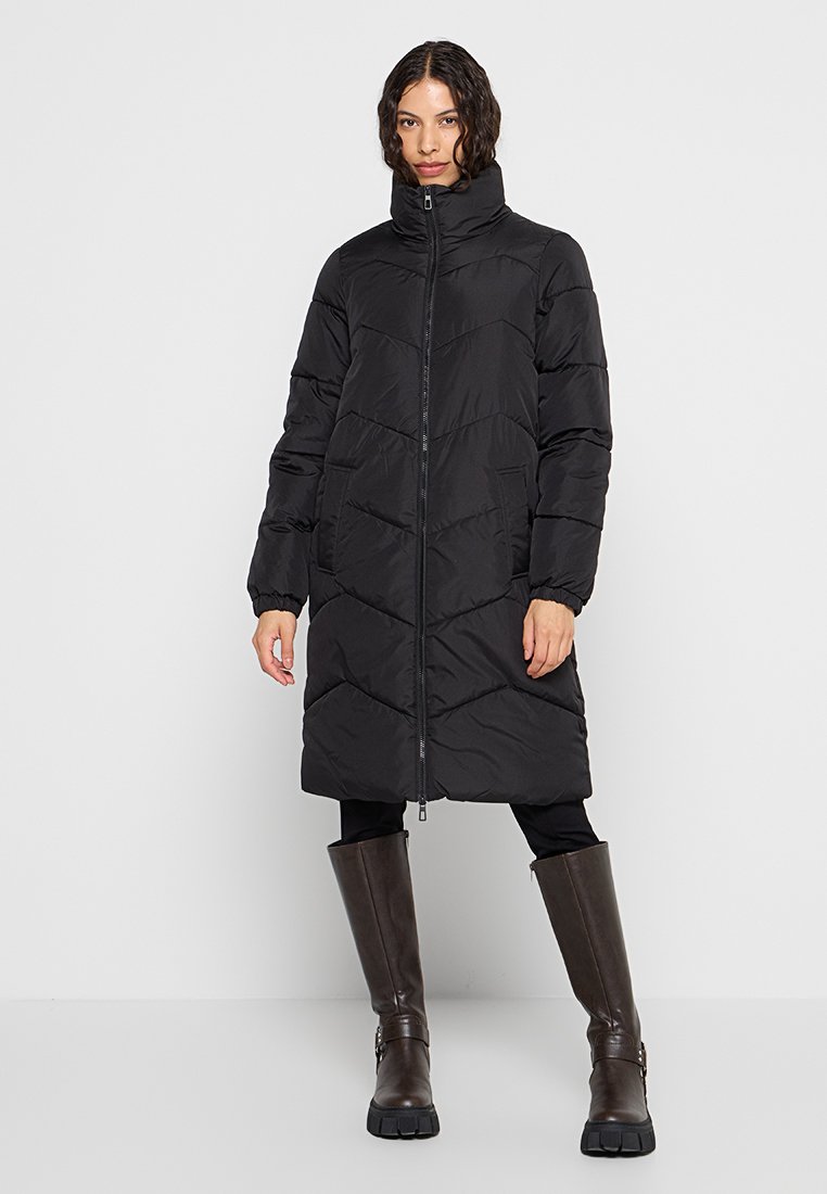 Schwarze Puffermantel mit hohem Kragen, Zickzack-Nähten und Frontreißverschluss. Mit langen Ärmeln und Taschen ausgestattet. Getragen über enganliegenden schwarzen Leggings und Stiefeln.