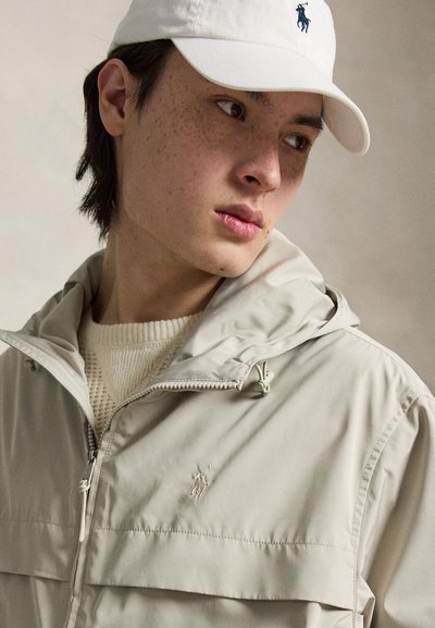 Jeune personne avec des taches de rousseur portant une veste beige zippée et un pull en maille crème, regardant sur le côté, avec une casquette de baseball blanche.