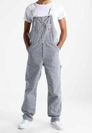 Homme portant une salopette rayée grise et blanche avec un t-shirt blanc et des baskets blanches, debout les mains dans les poches.