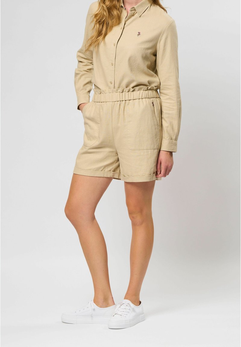 Beige, lichtgewicht, langemouwhemd met een knoopsluiting aan de voorkant en shorts met een elastische tailleband, gecombineerd met witte sneakers.